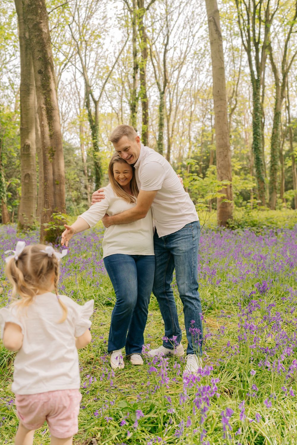 A family poses in a bluebell woodland, with a toddler running toward her embracing parents among purple wildflowers.