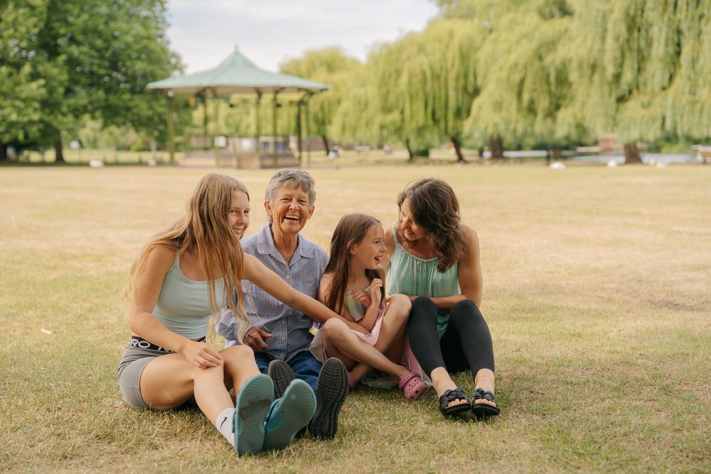 Four females sitting together on green grass in a sunny park, laughing and enjoying time together near a bandstand.