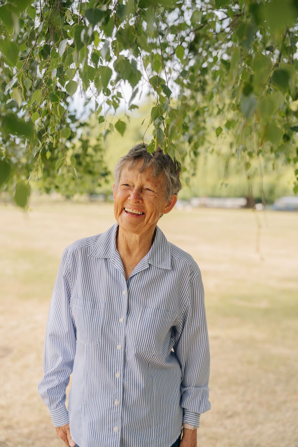 Smiling senior woman with short gray hair wearing a blue striped shirt standing outdoors under birch tree branches.