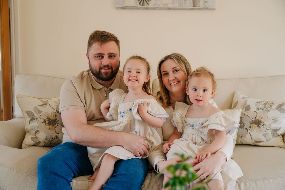A happy family of four poses on a cream sofa, parents holding two young girls in matching white floral dresses.