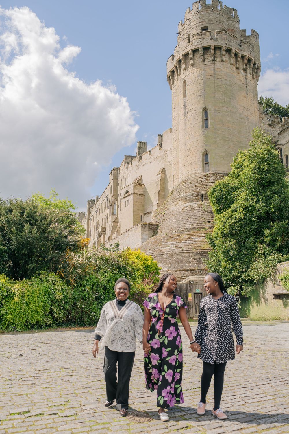 Three women walking on cobblestone path in front of historic Warwick Castle on a sunny day.