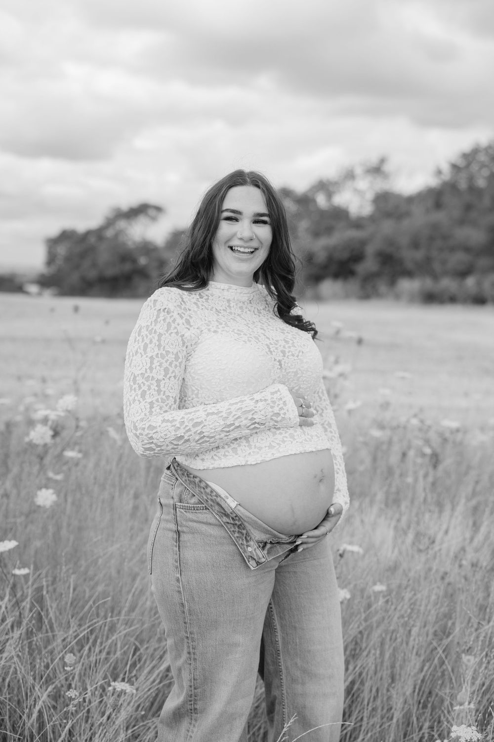 Black and white maternity photo of smiling pregnant woman in lace top and jeans standing in a wildflower field.