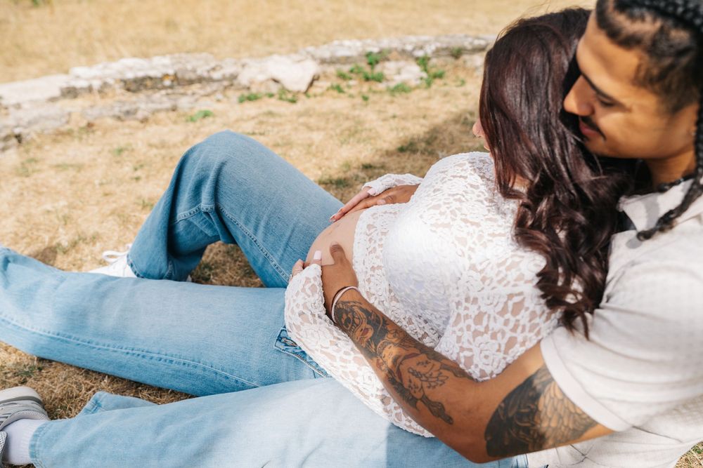 Couple sitting outdoors, man with tattoos embracing pregnant woman in white lace top and jeans.