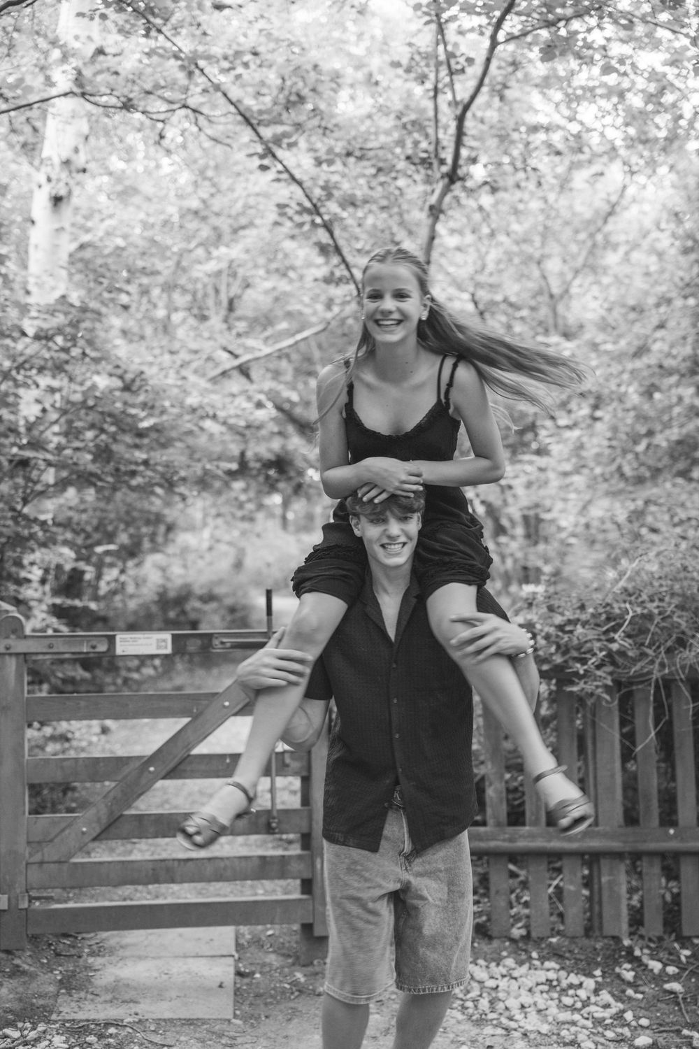 Black and white photo of a smiling girl sitting on a young man's shoulders near a wooden gate in a forest.