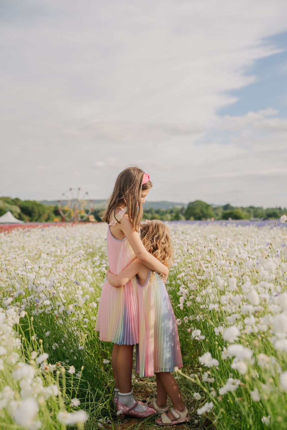 Two young girls in rainbow dresses embrace in a white wildflower field under a cloudy sky.