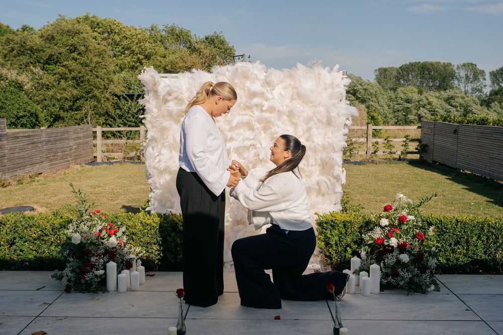 Same-sex couple proposal outdoors, one partner kneeling with flowers, white floral backdrop, garden setting. warwickshire wedding photograph