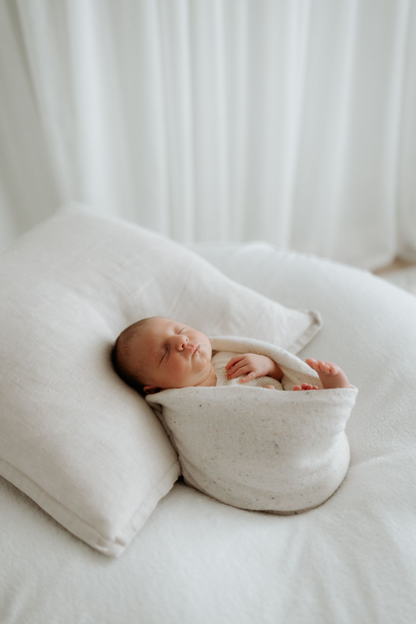 Newborn baby wrapped in white blanket rests peacefully on soft white surface.