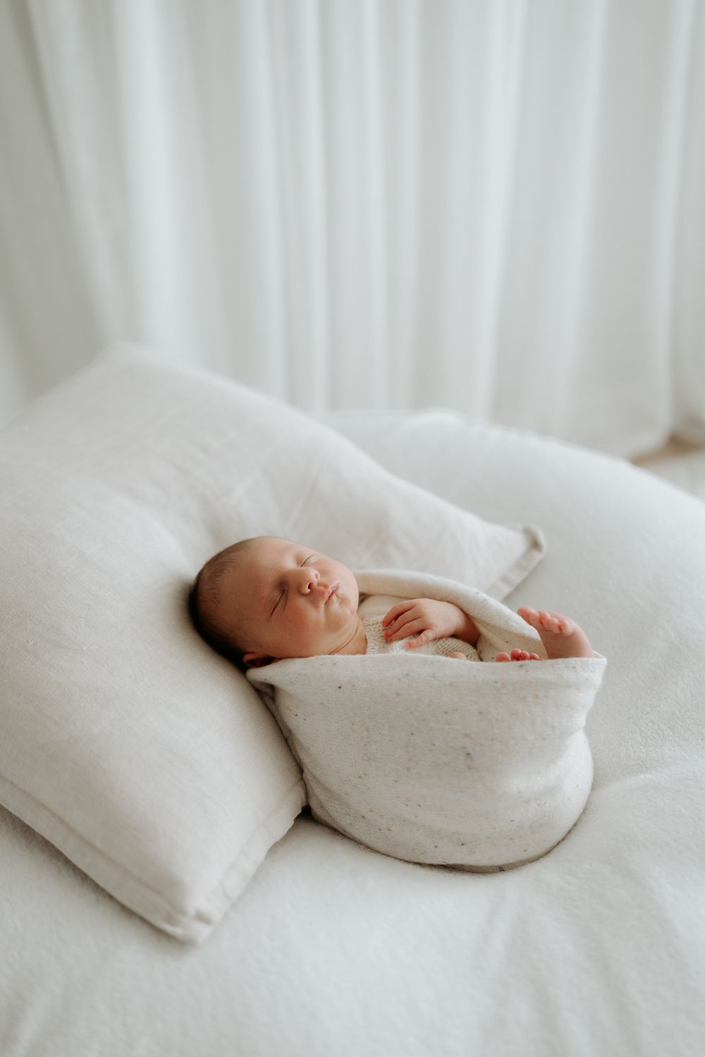 Newborn baby wrapped in white blanket rests peacefully on soft white surface.