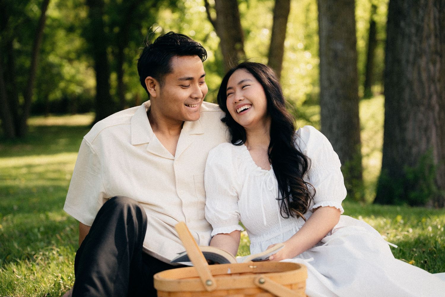 Happy Asian couple laughing together at a sunny park picnic with a wicker basket nearby.