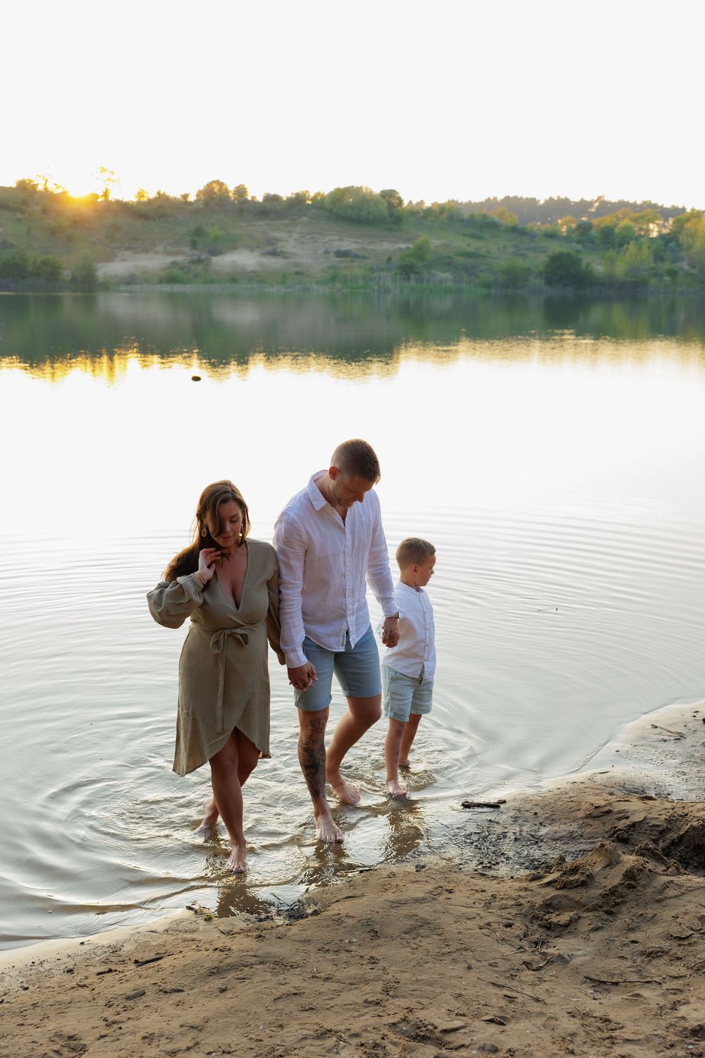 Family of three wading in shallow lake water at golden sunset, parents and child enjoying summer day outdoors.