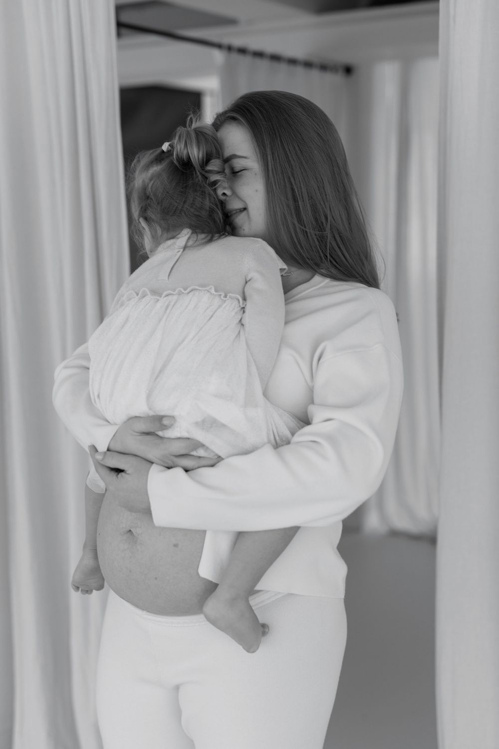 Black and white photo of a pregnant woman in white outfit tenderly holding and hugging a young child indoors.