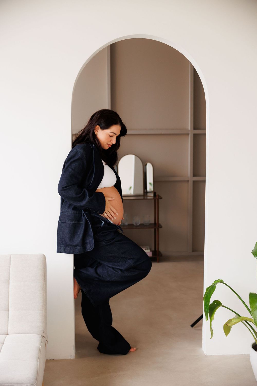 Pregnant woman in navy suit cradling baby bump, standing in elegant white arched doorway of modern home.