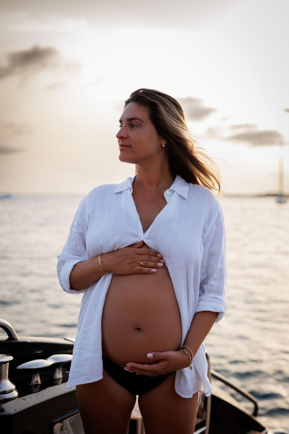 Pregnant woman in white shirt and bikini cradling her baby bump on a boat at sunset over the ocean.