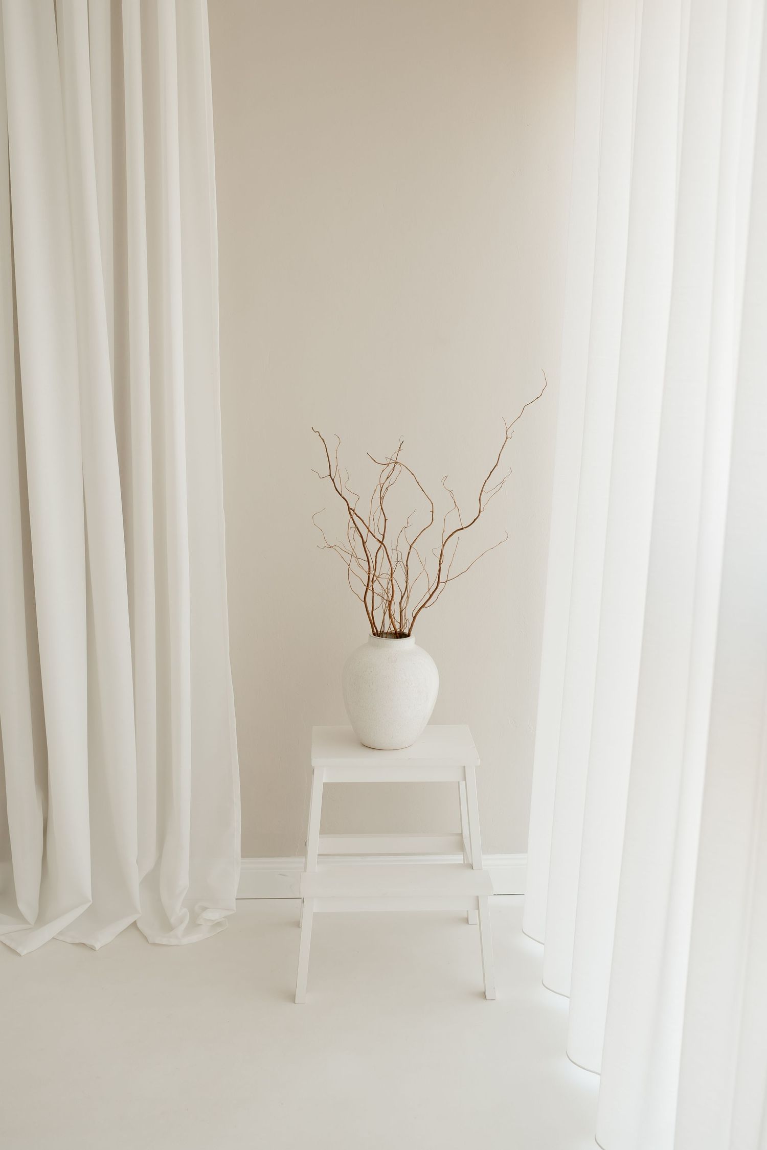 Minimalist white interior with white curtains and a white vase containing curvy twigs on a stepped pedestal.
