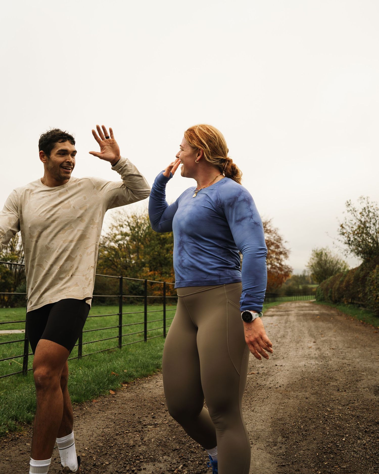 Two people in athletic wear sharing a playful moment while exercising on a dirt path surrounded by autumn trees.