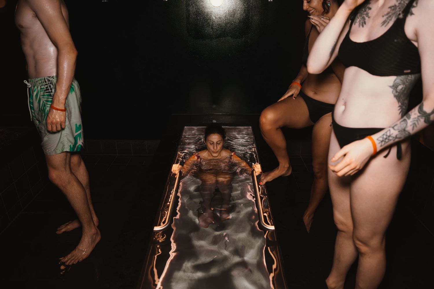 A person lies on a skateboard illuminated by neon lights while others gather around in swimwear at night.