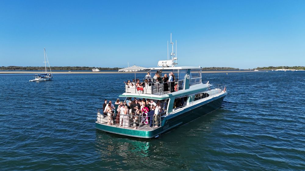 A party cruise boat filled with guests sailing on calm blue waters under a clear sunny sky.