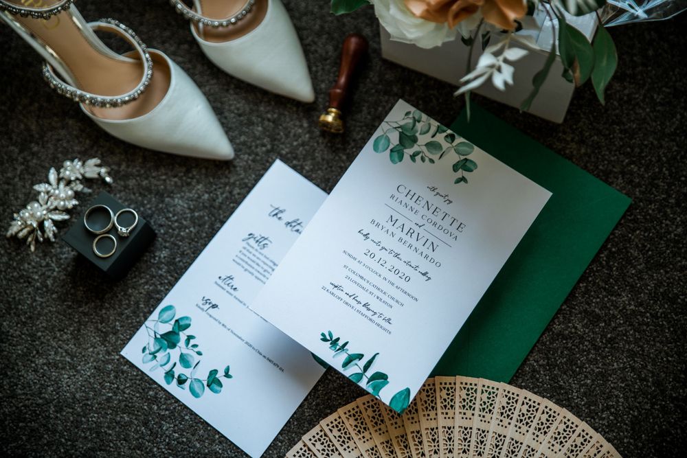 Wedding invitation with green floral design displayed alongside bridal shoes and rings.