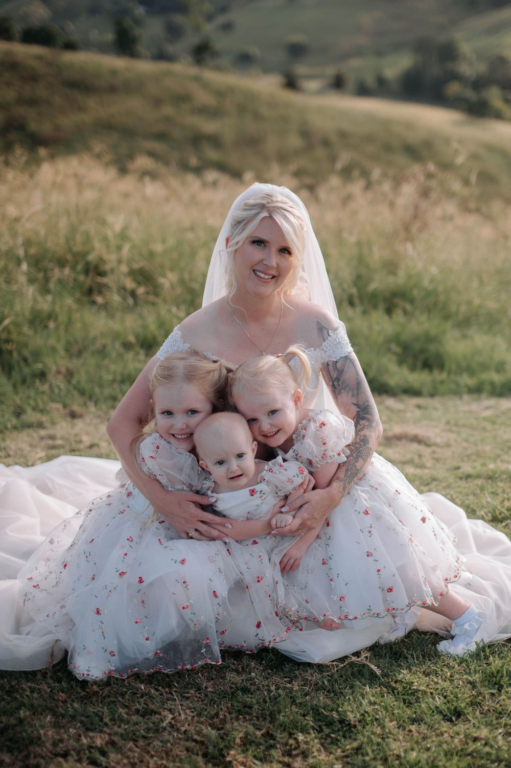 A bride in a white dress embraces three children wearing matching outfits in a grassy field.