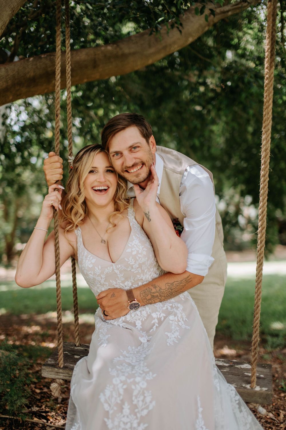 A joyful wedding couple laughs together on a rustic rope swing under a tree during an outdoor photoshoot.