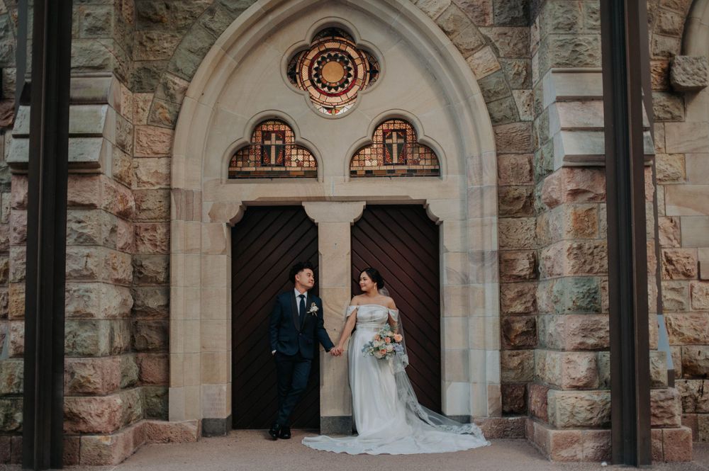 A wedding couple stands in a gothic stone church doorway with arched details.