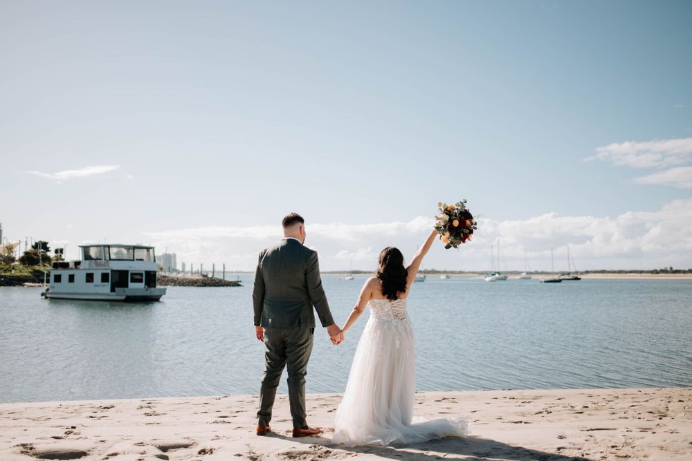 Newlyweds hold hands while standing on a beach with boats in the harbor on their wedding day.