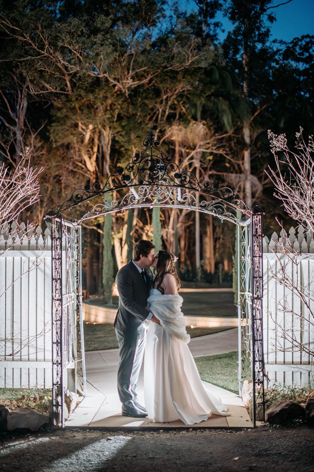 Romantic nighttime wedding photo under an illuminated garden archway.