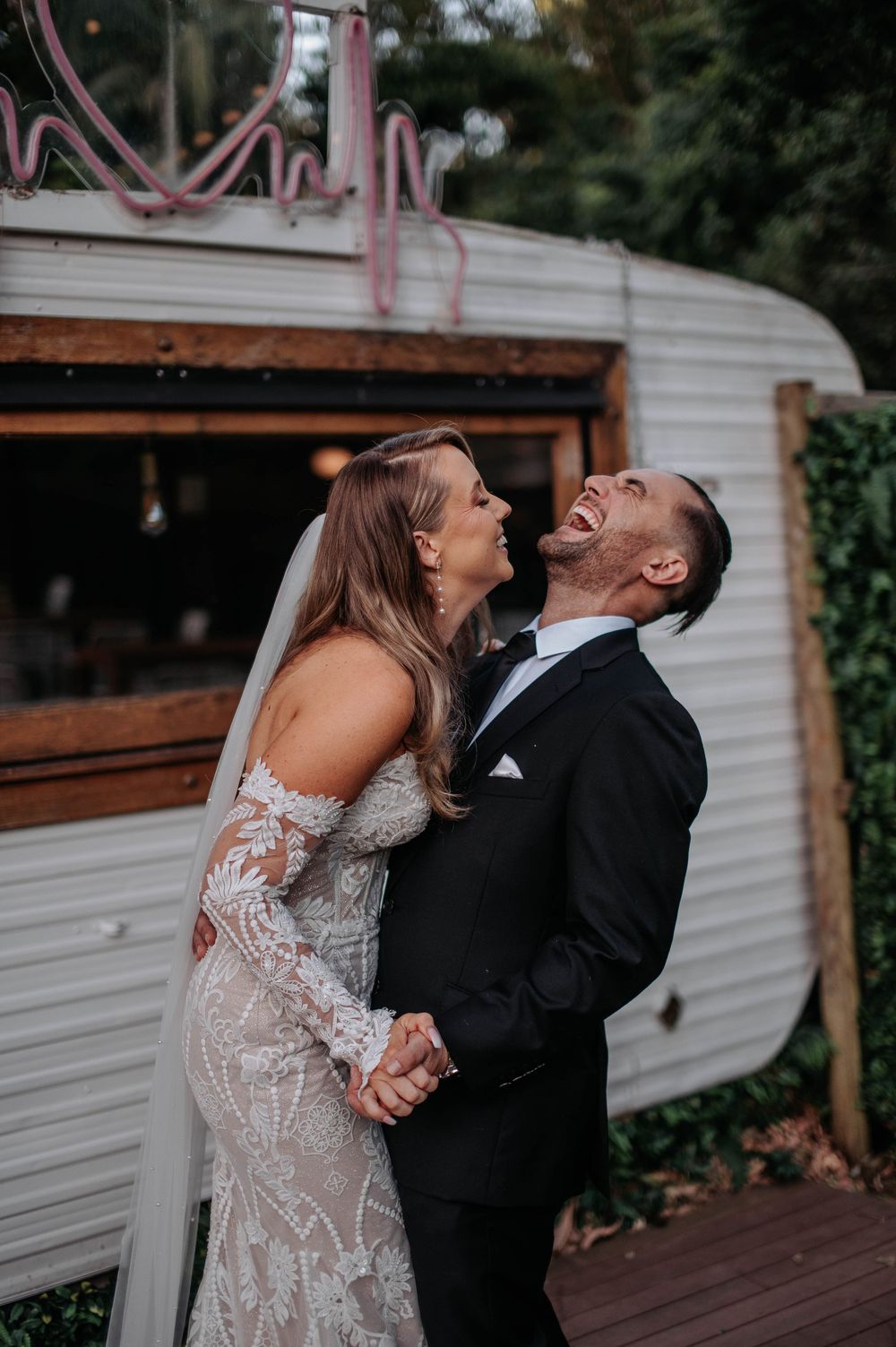 A couple shares a joyful laughing moment together in front of a vintage white camper trailer on their wedding day.