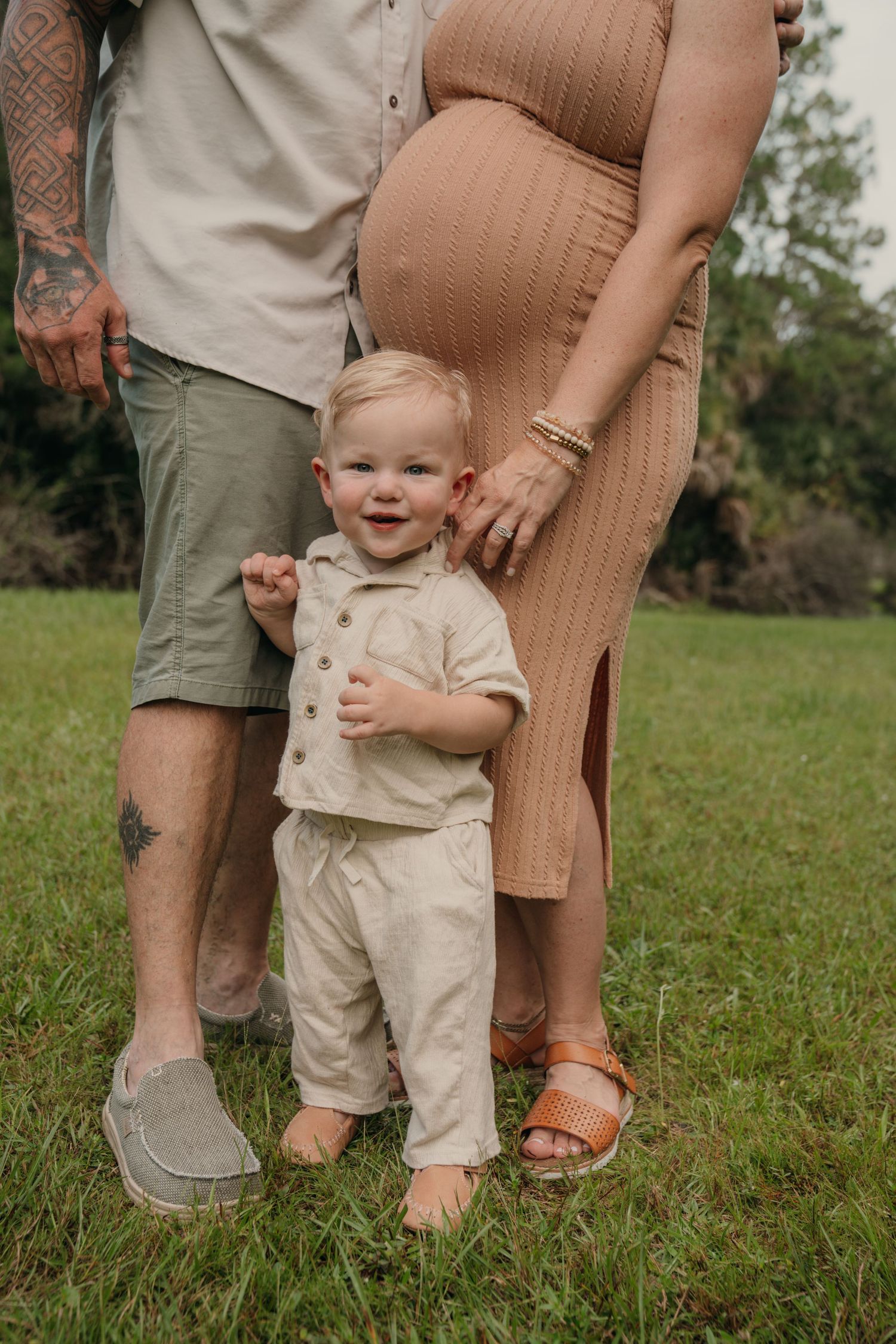 Smiling toddler stands between tattooed father and pregnant mother in neutral tones outdoors on green grass.