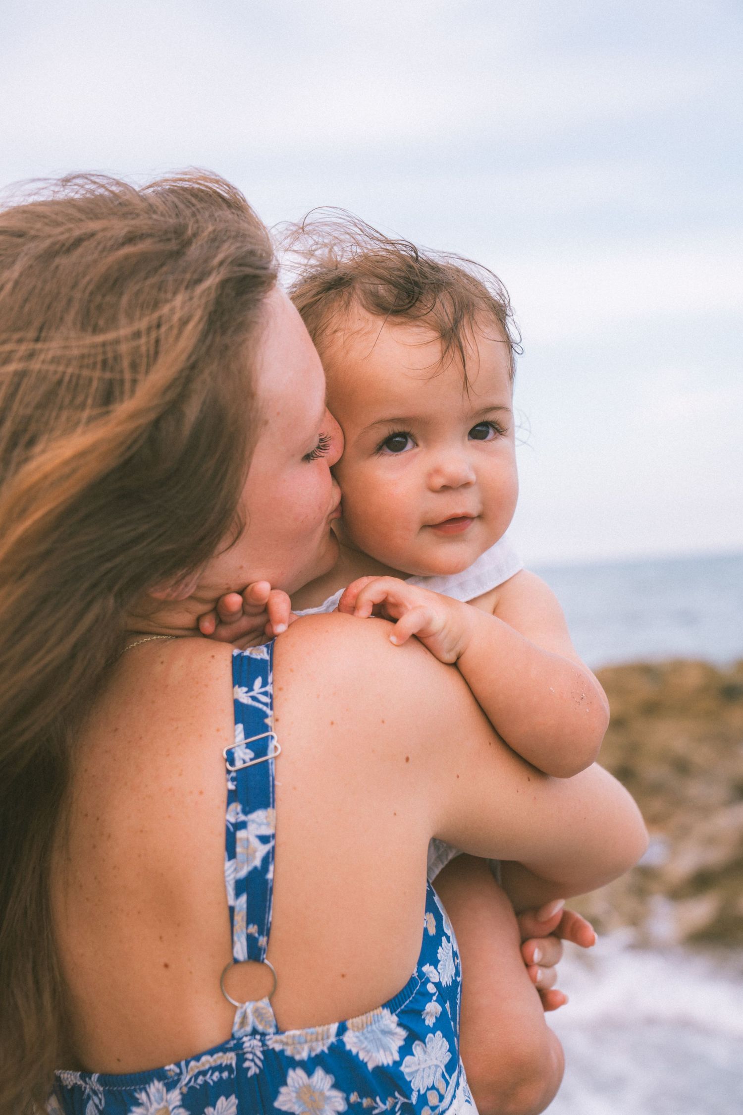 A mother in a blue floral swimsuit kisses her baby on the cheek at the beach.