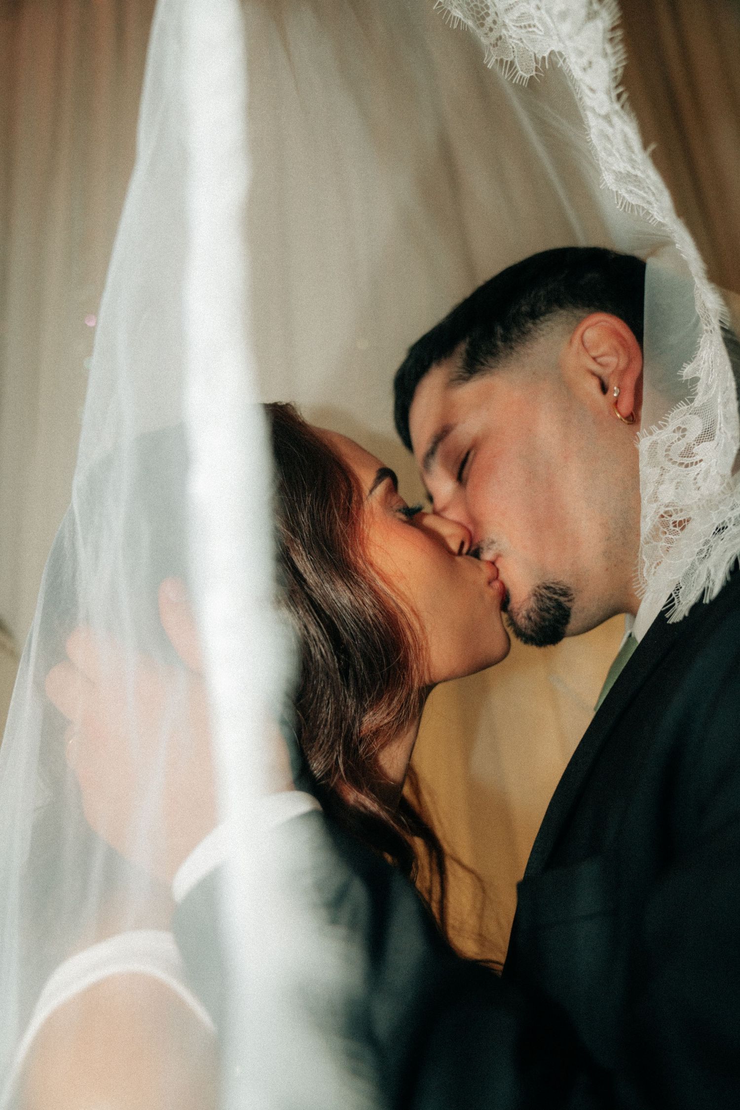 A bride and groom share a romantic kiss beneath a flowing white wedding veil in soft natural light.