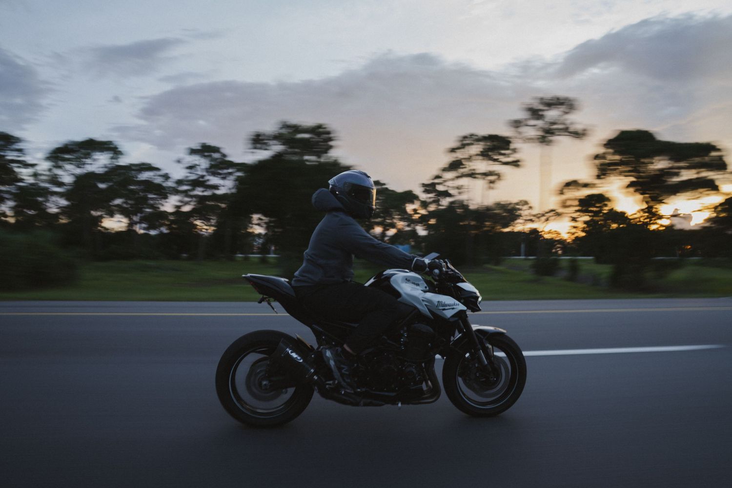 Motorcyclist riding a sport bike on an open road at dusk, with trees and cloudy sky in the background.