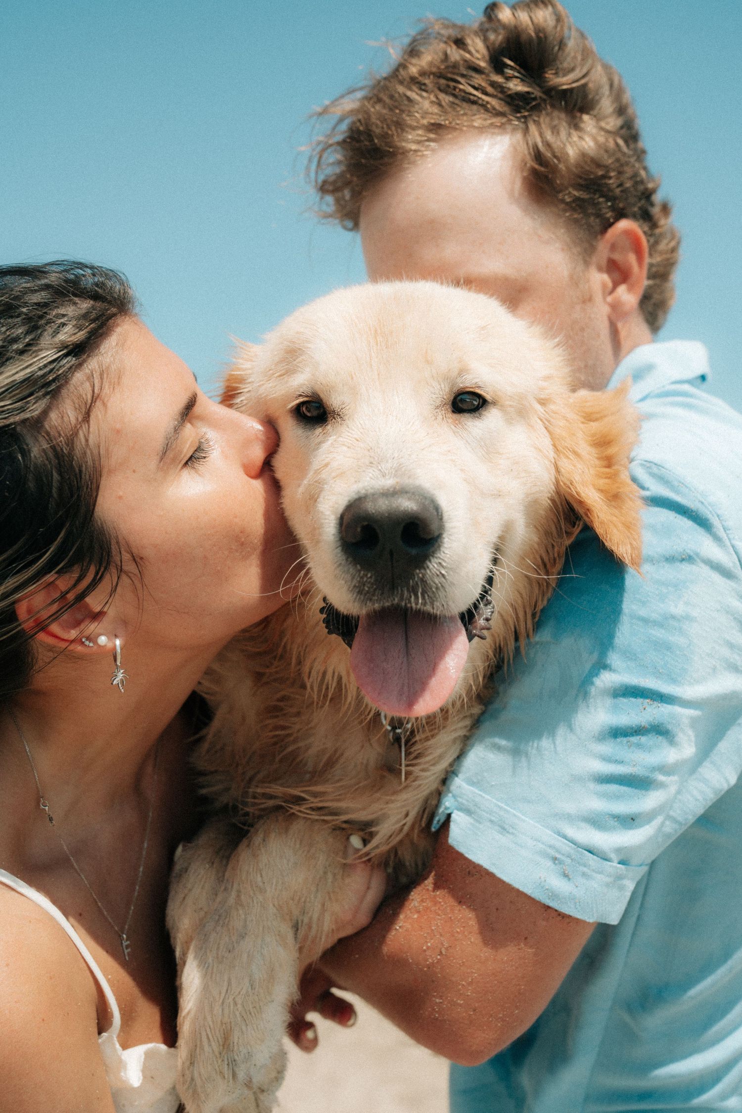 A happy couple kissing their wet golden retriever dog on the beach under a clear blue sky.