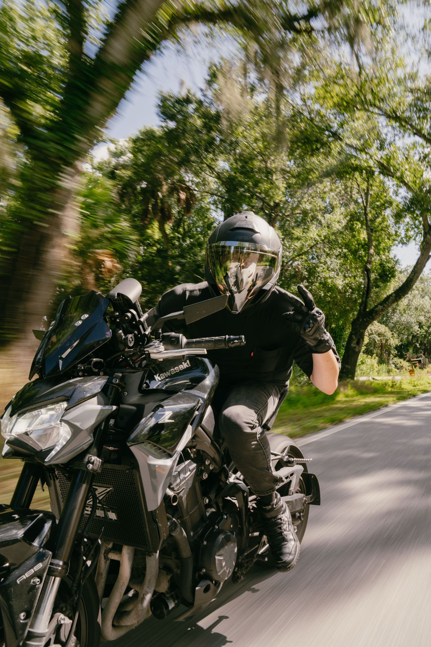 Motorcyclist in black gear and helmet rides a dark sport motorcycle at speed on a tree-lined road.