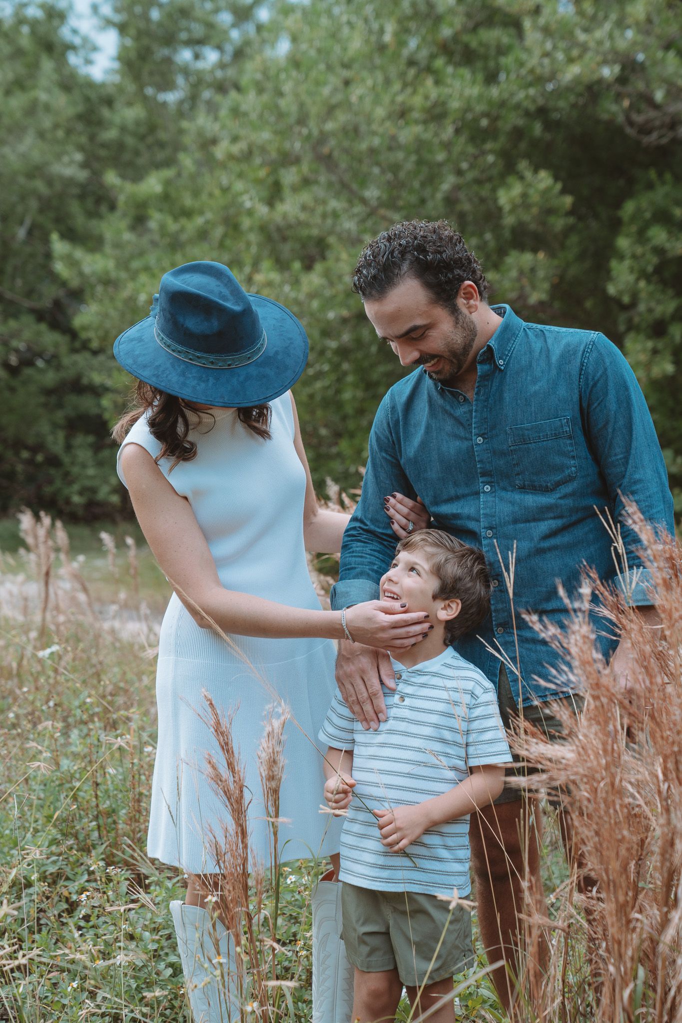 A family shares a tender moment together outdoors among tall grass and greenery on a summer day.
