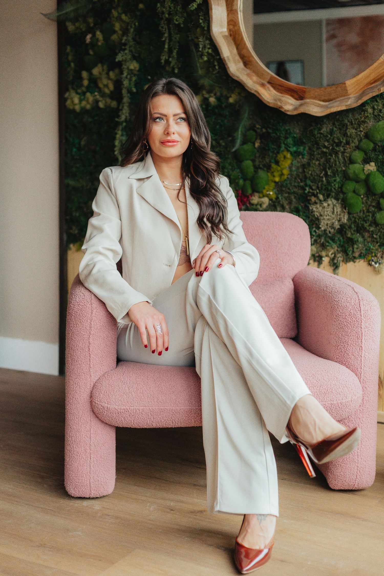 Elegant woman in cream pantsuit posing in a pink armchair with green wall backdrop.