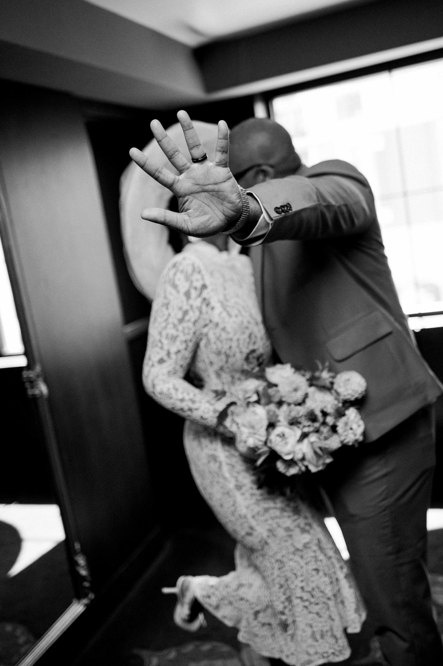 A black and white candid wedding photo showing a couple dancing, with the bride in lace and holding a floral bouquet.