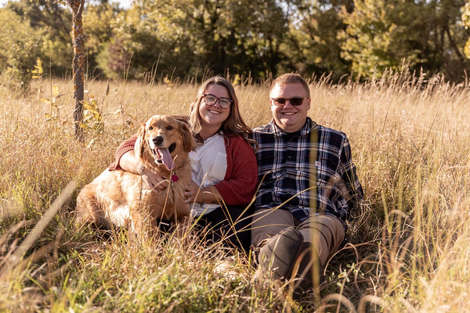Couple smiling and sitting in tall golden grass with their golden retriever dog on a sunny autumn day.