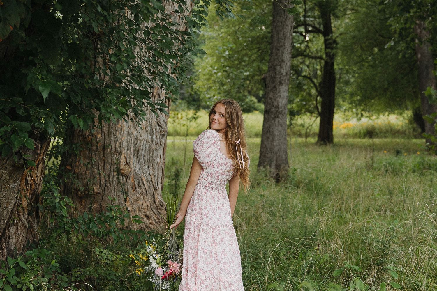Woman in floral dress walking through sunlit wooded area.