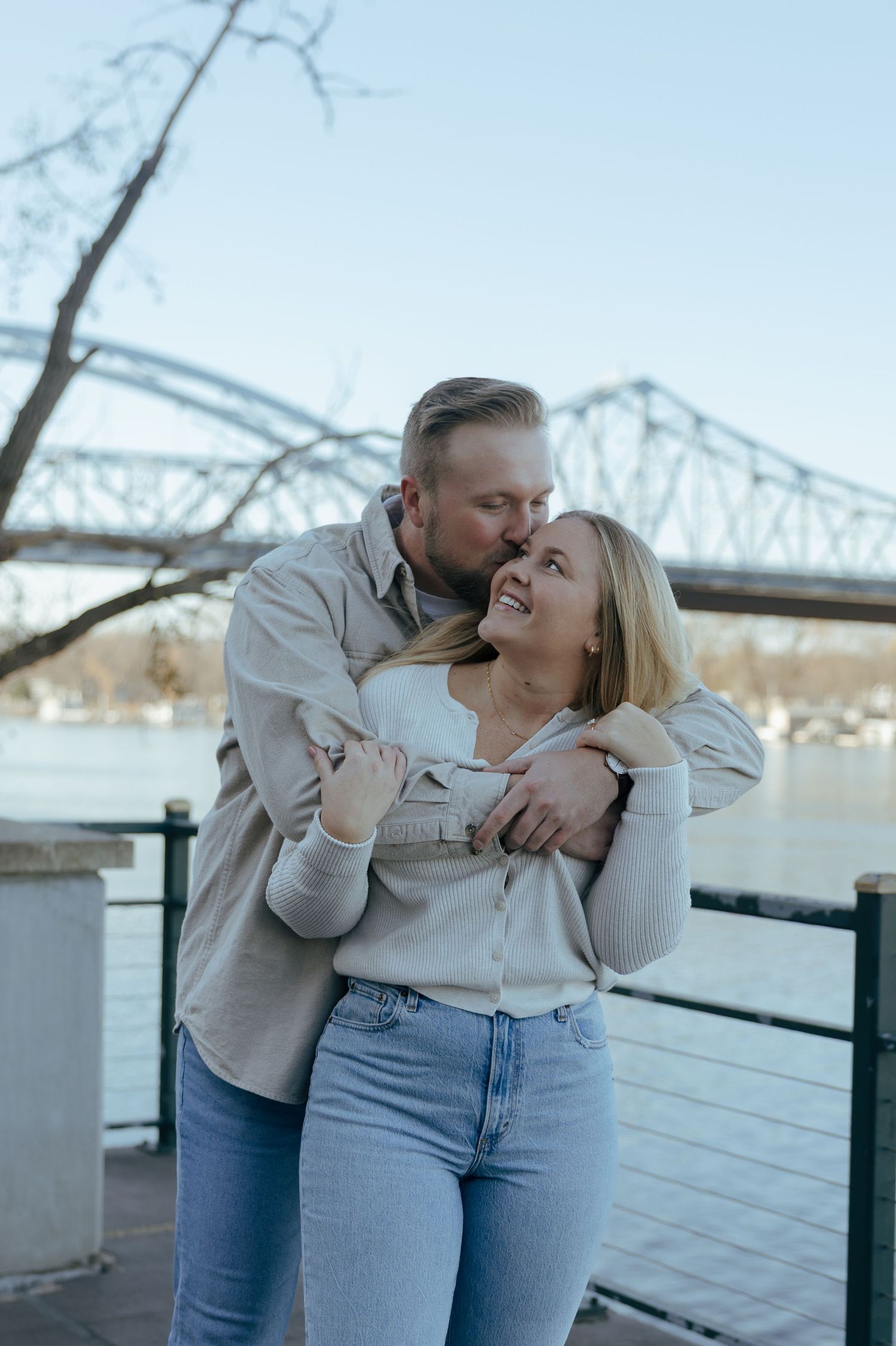 A couple shares a romantic moment by the river with bridge architecture in background.
