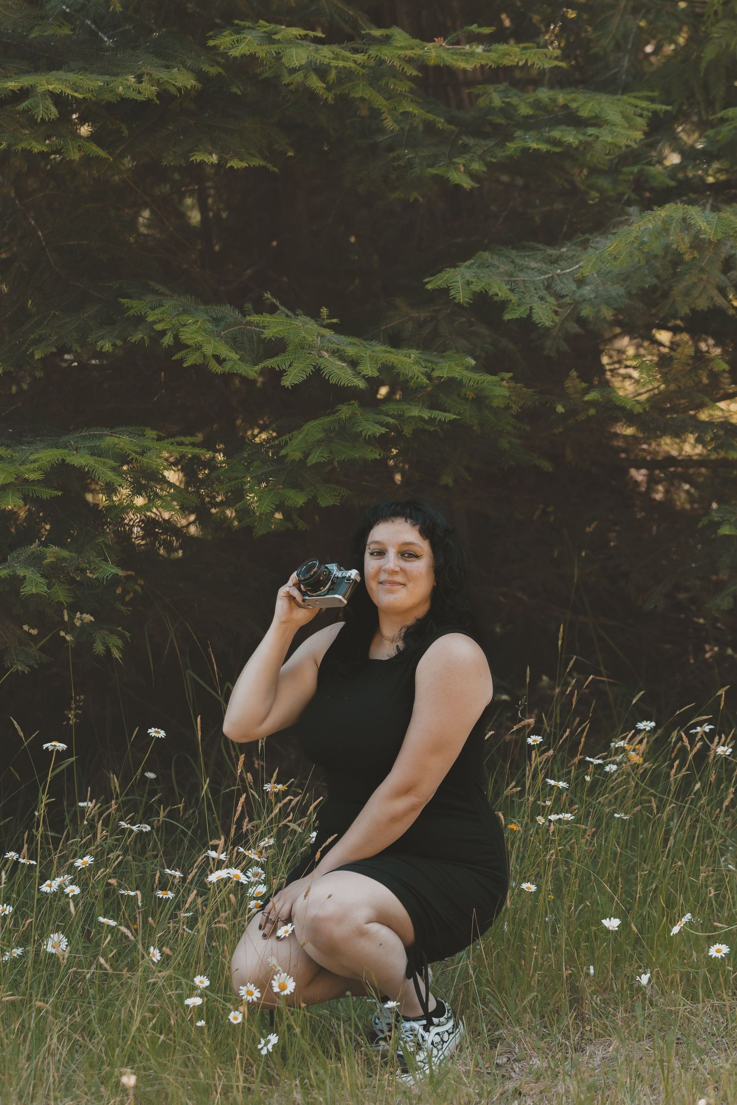 A woman crouched is in a field of daisys in front of a tree holding a camera.