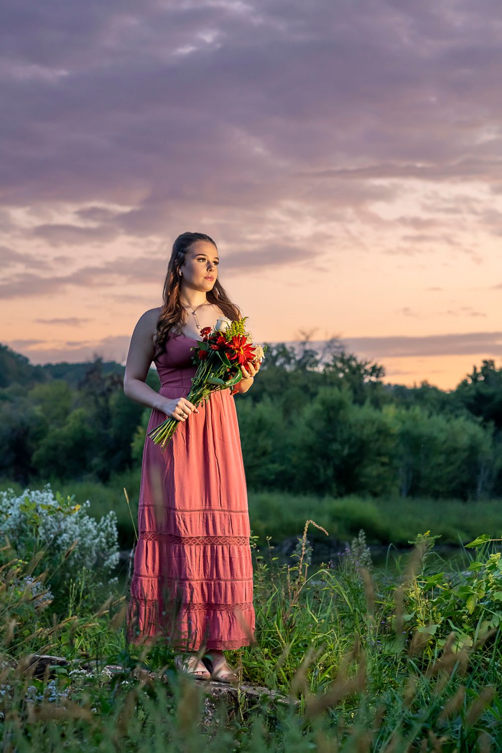 A woman in a pink flowy dress gathers colorful wildflowers in a meadow at sunset with dramatic clouds overhead.