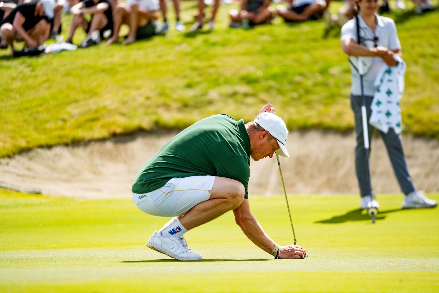 Profesjonell golfspiller i grønn skjorte gjør seg klar til å putte på greenen, mens en tilskuer følger med fra banen under turneringsspill.