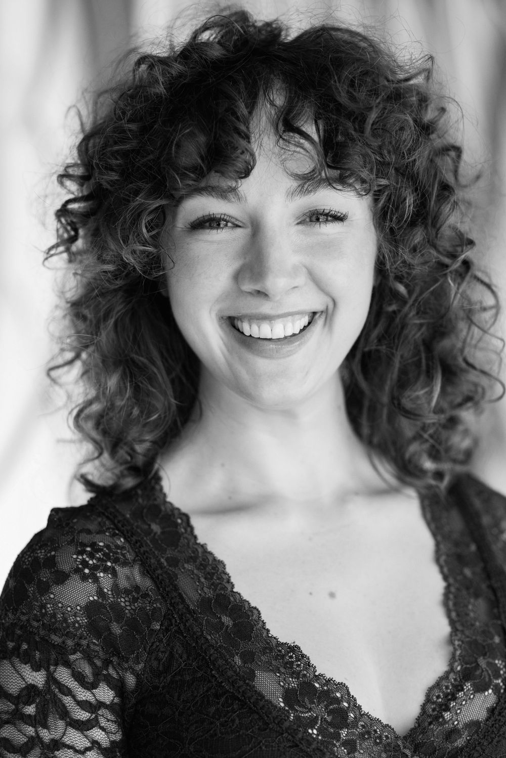 Black and white portrait of a smiling person with dark curly hair wearing a black lace top.