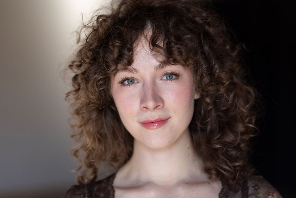 Someone with natural curly brown hair and a gentle smile poses against a dark background in this headshot portrait.