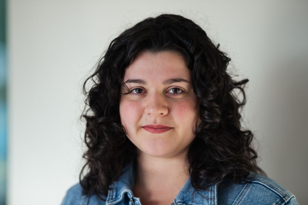A person with dark curly hair and a warm smile wearing a denim jacket against a light background.