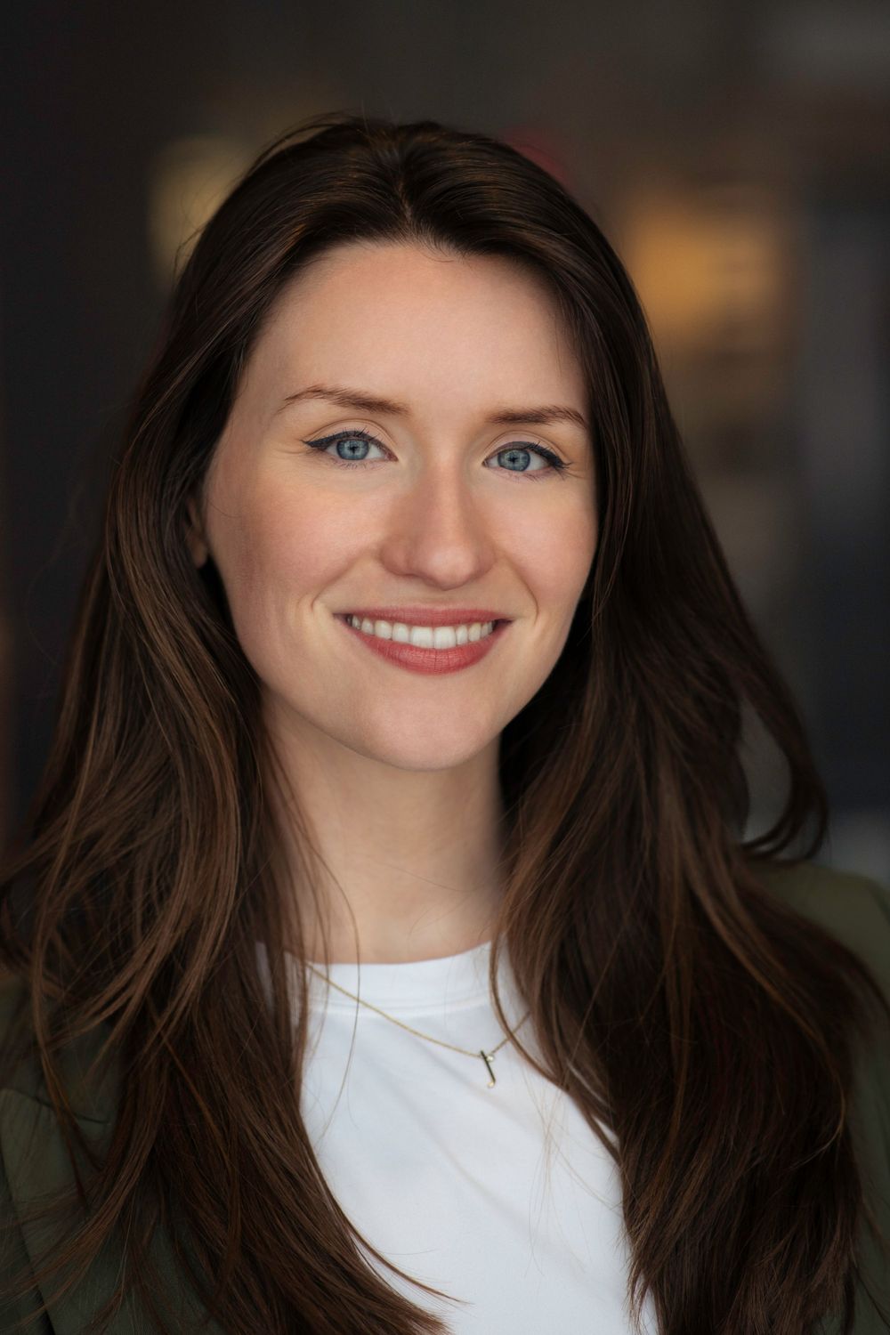 Professional headshot of a person smiling at the camera wearing a white top and delicate necklace against a blurred background.