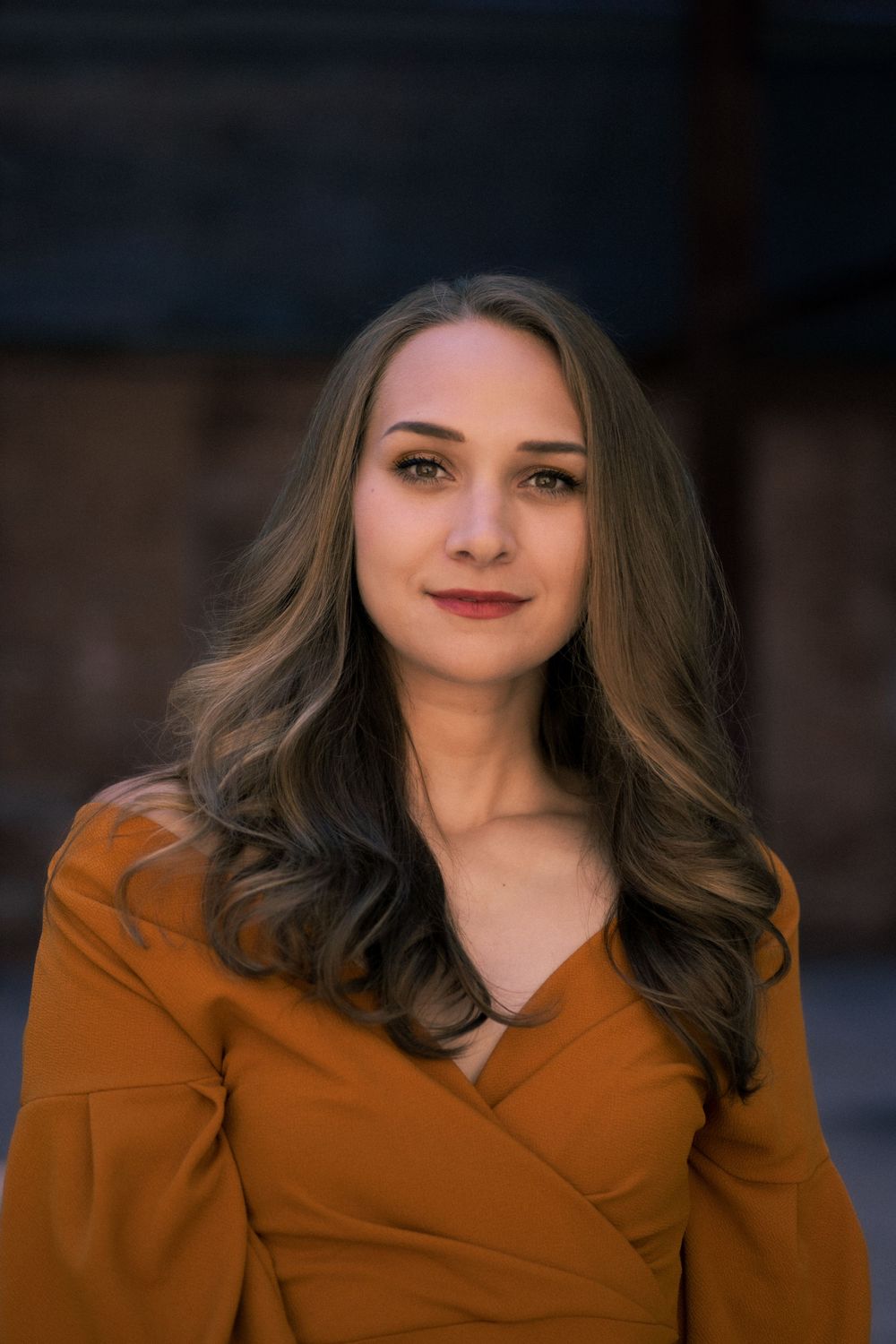 Portrait featuring warm tones with a burnt orange wrap top and flowing brown hair against a soft blurred background.