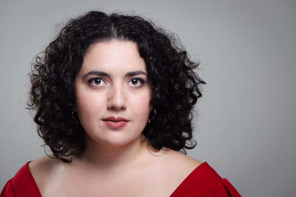 A striking studio portrait with dark curls and a red shirt against a gray background.