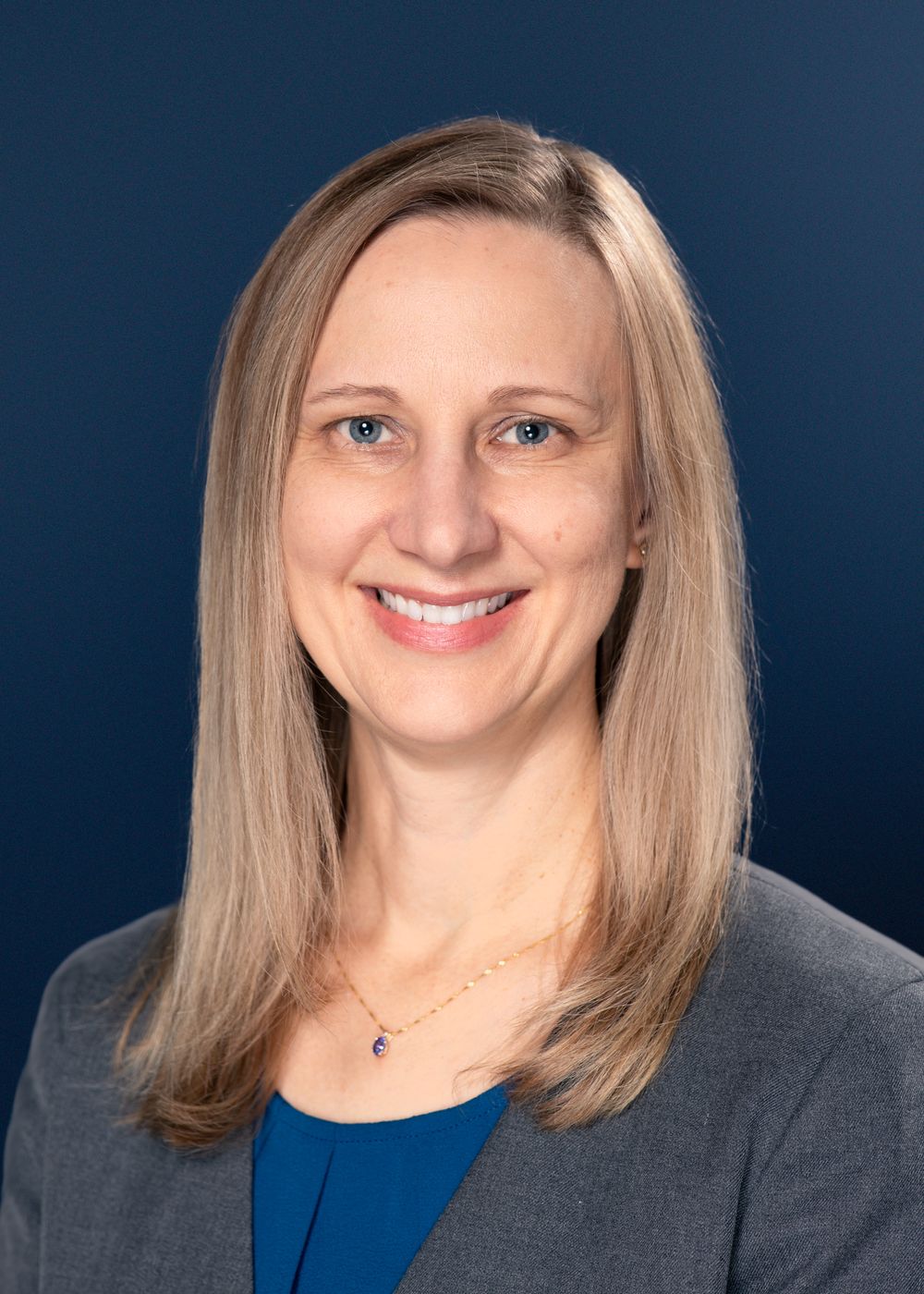 Professional headshot of a person with blonde shoulder-length hair wearing a gray blazer and blue top against navy background.